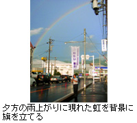 写真：夕方の雨上がりに現れた虹を背景に旗を立てる